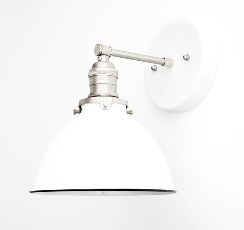 Puede incluir: Un aplique de pared con una pantalla de metal blanca en forma de c&uacute;pula, un casquillo de n&iacute;quel cepillado y una placa de pared circular blanca a juego. El brazo conecta la pantalla a la base con una bisagra visible.
