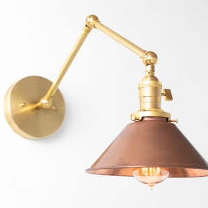 May include: A wall-mounted brass and copper-toned lamp with an adjustable arm. The lamp features a circular base, a pivoting arm, and a conical shade. The light bulb is visible within the shade.