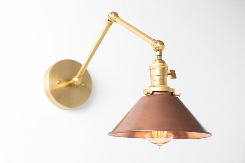 May include: A wall-mounted brass and copper-toned lamp with an adjustable arm. The lamp features a circular base, a pivoting arm, and a conical shade. The light bulb is visible within the shade.