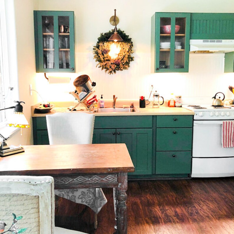 May include: A kitchen with green cabinets, a wooden table, and a white chair. A wreath hangs above the sink, and a stove and refrigerator are visible. The room has a warm, inviting atmosphere.