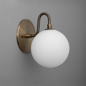 May include: A wall-mounted light fixture with a round, frosted white glass globe. The fixture has a curved, antique brass arm and a circular backplate. The design is simple and elegant, suitable for various interior styles.