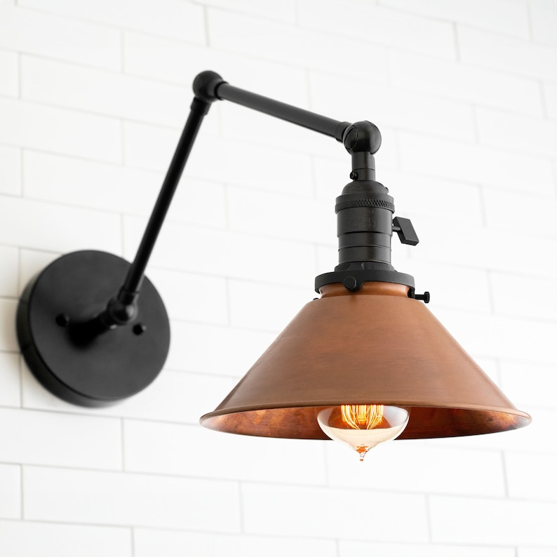 May include: A black wall-mounted light fixture with a copper-colored shade. The fixture has an adjustable arm and a round base. The light bulb is visible inside the shade. The fixture is mounted on a white brick wall.