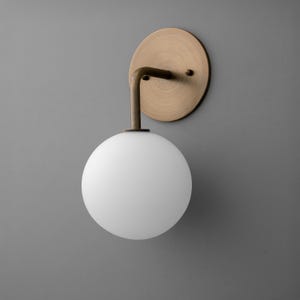 May include: A modern wall sconce featuring a round, white globe light. The light is supported by a curved, dark metal arm that connects to a circular, brass-colored wall plate. The design is minimalist and elegant.