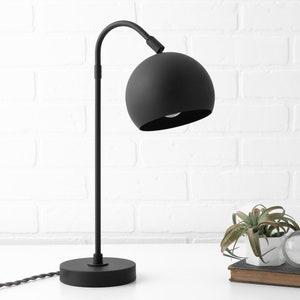 May include: A black table lamp with a curved arm and a round, black shade. The lamp is on a white surface with a small plant and a book.