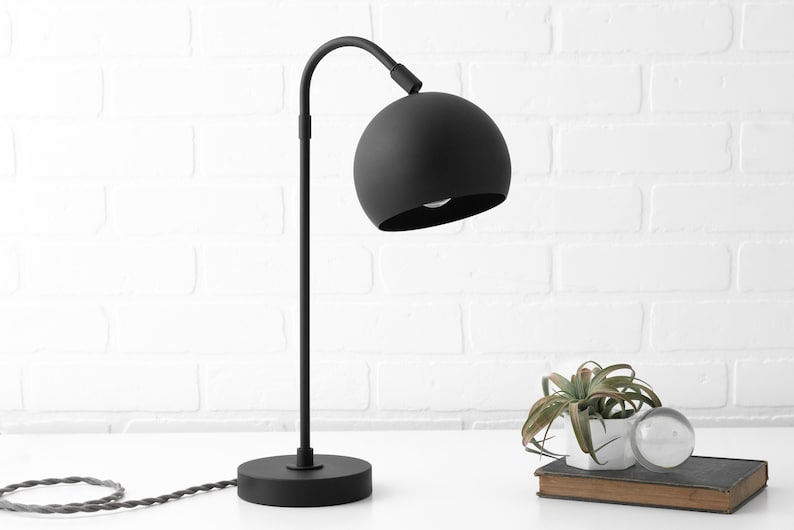May include: A black table lamp with a curved arm and a round, black shade. The lamp is on a white surface with a small plant and a book.