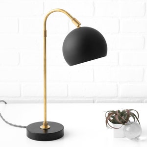 May include: A black and gold table lamp with a round black shade. The lamp has a curved arm and a black base.