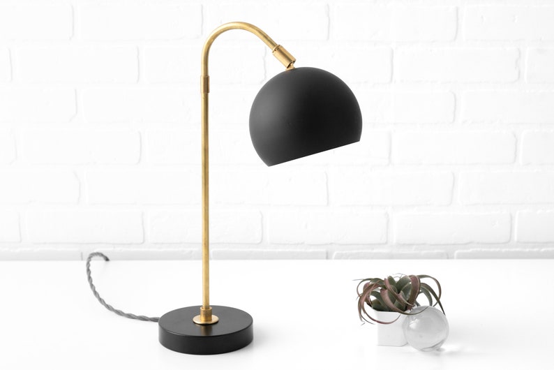 May include: A black and gold table lamp with a round black shade. The lamp has a curved arm and a black base.