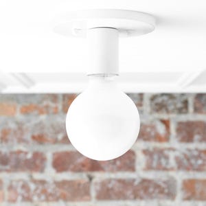 May include: A white, modern flush mount ceiling light fixture with a large, round bulb. The fixture is mounted on a white ceiling with decorative molding. The background features a blurred brick wall.