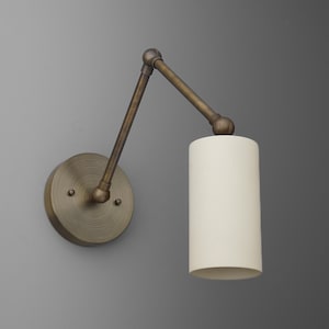 May include: A wall-mounted lamp with a cylindrical off-white shade and an adjustable arm. The lamp has a circular base and arm in a bronze finish. The design is simple and modern, suitable for various interior styles.