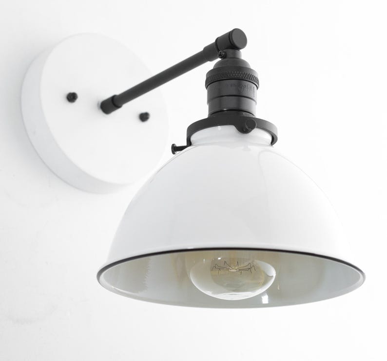 Puede incluir: Un aplique de pared con base circular blanca, brazo de metal negro y casquillo negro. La l&aacute;mpara incluye una pantalla blanca ancha en forma de c&uacute;pula con borde negro y una bombilla de cristal transparente en el centro.