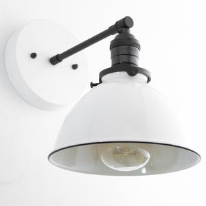 May include: A wall-mounted light fixture featuring a white circular base, a black metal arm, and a black socket. The fixture includes a wide, white dome-shaped shade with a black rim and a single clear glass light bulb centered inside.
