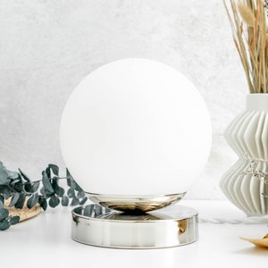 May include: A spherical white glass lamp shade sits atop a polished silver-toned circular metal base. The lamp features a smooth, opaque white globe design that rests securely on the reflective metallic stand, creating a clean and balanced appearance.