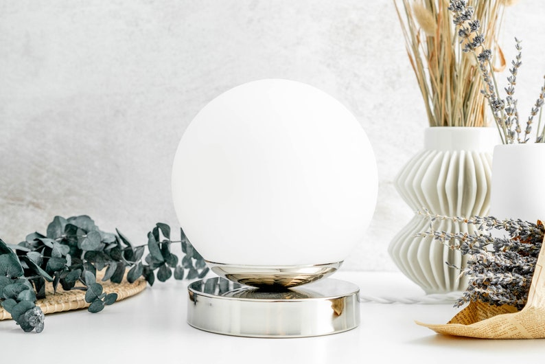 May include: A spherical white glass lamp shade sits atop a polished silver-toned circular metal base. The lamp features a smooth, opaque white globe design that rests securely on the reflective metallic stand, creating a clean and balanced appearance.