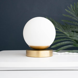 May include: A table lamp featuring a spherical white glass globe shade resting on a circular brushed brass base. The lamp includes a white twisted fabric power cord extending from the side of the metallic base, providing a clean and simple design.