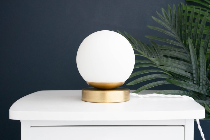 May include: A table lamp featuring a spherical white glass globe shade resting on a circular brushed brass base. The lamp includes a white twisted fabric power cord extending from the side of the metallic base, providing a clean and simple design.