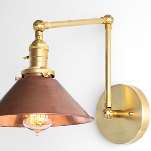 May include: A wall-mounted, adjustable lamp with a copper-colored shade and brass-colored arm and base. The lamp features an exposed bulb and a conical shade. The design is industrial and vintage.