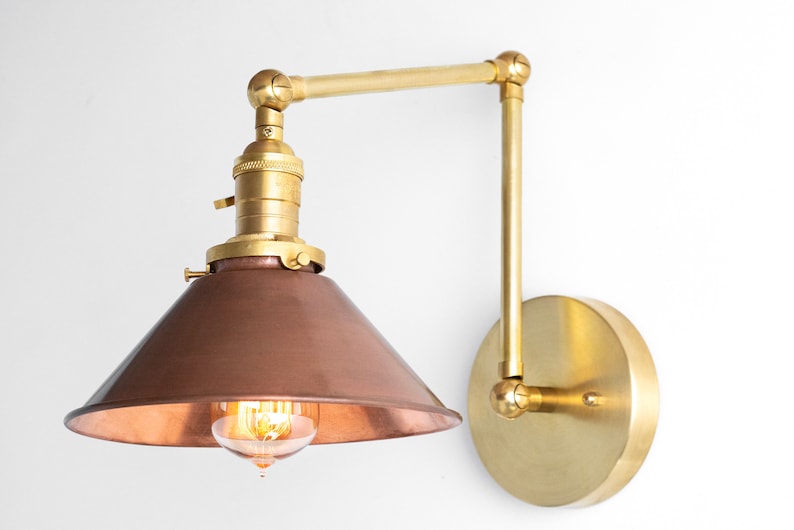 May include: A wall-mounted, adjustable lamp with a copper-colored shade and brass-colored arm and base. The lamp features an exposed bulb and a conical shade. The design is industrial and vintage.