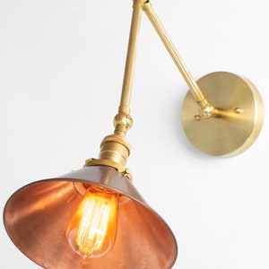 May include: A brass and copper wall-mounted light fixture with an exposed bulb. The fixture has an adjustable arm and a conical shade. The light is on, illuminating the interior of the shade.