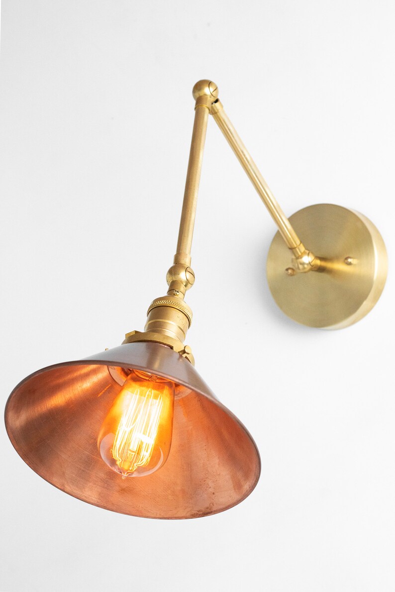 May include: A brass and copper wall-mounted light fixture with an exposed bulb. The fixture has an adjustable arm and a conical shade. The light is on, illuminating the interior of the shade.