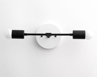 May include: A two-light wall sconce featuring a circular white mounting plate and a horizontal black metal bar. Two cylindrical black sockets are positioned at each end of the bar, each holding a round white light bulb, creating a linear design.