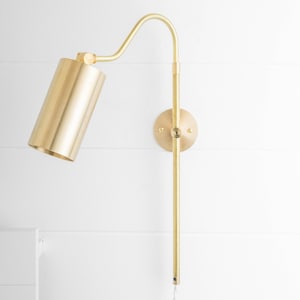 May include: A gold-toned wall-mounted light fixture with a cylindrical shade and a long, adjustable arm. The light is designed to be used as a reading lamp or task light.