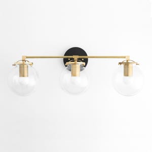 May include: A three-light vanity fixture with clear globe shades and brass-colored accents. The fixture is mounted on a black circular backplate. The horizontal bar and light fixtures are brass-colored.