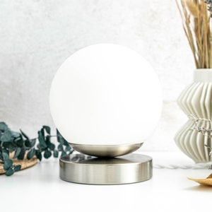 May include: A table lamp featuring a spherical white glass shade resting on a circular brushed metal base. The lamp has a smooth, rounded design with a metallic finish on the bottom portion and a clean, opaque white glass globe on top.