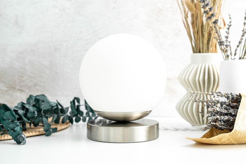 May include: A table lamp featuring a spherical white glass shade resting on a circular brushed metal base. The lamp has a smooth, rounded design with a metallic finish on the bottom portion and a clean, opaque white glass globe on top.