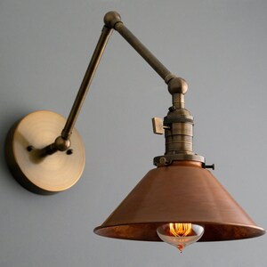 May include: Vintage-style wall sconce with a bronze finish. The lamp features an adjustable arm and a copper-colored shade. The exposed bulb emits a warm glow, adding a touch of industrial design to any space.