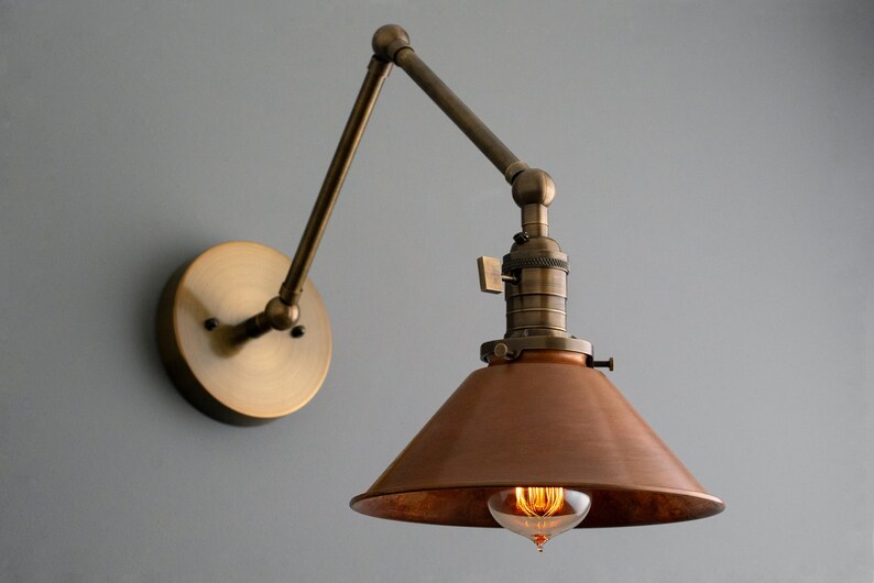 May include: Vintage-style wall sconce with a bronze finish. The lamp features an adjustable arm and a copper-colored shade. The exposed bulb emits a warm glow, adding a touch of industrial design to any space.