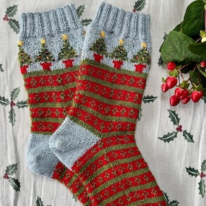 Peut inclure: Une paire de chaussettes de No&euml;l tricot&eacute;es. Les chaussettes pr&eacute;sentent un bord et un bout du pied bleu clair, avec un motif ray&eacute; rouge et vert sur le corps. Le bord pr&eacute;sente un motif de sapins de No&euml;l en pots.