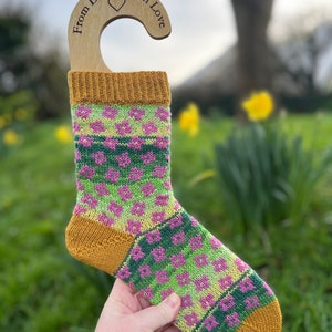 May include: A pair of colorful knitted socks with a floral pattern in shades of pink, green, and yellow. The socks are hanging on a wooden sock hanger with the text "From Devon With Love".