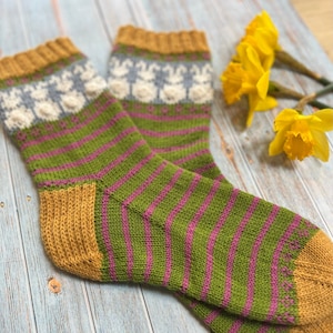 May include: A pair of knitted socks with a green and pink striped pattern and a yellow cuff. The socks have a white and blue pattern with a bunny design at the top.