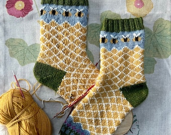 May include: A pair of hand-knitted socks with a honeycomb pattern in yellow and white, green and blue accents, and a bee motif. The socks are displayed on a wooden sock blocker.
