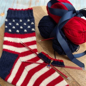 Pode incluir: Uma meia de tricô vermelha, branca e azul com um padrão de estrelas no topo e listras na parte inferior. A meia está sobre uma superfície de madeira com novelos de lã vermelha, branca e azul e agulhas de tricô.