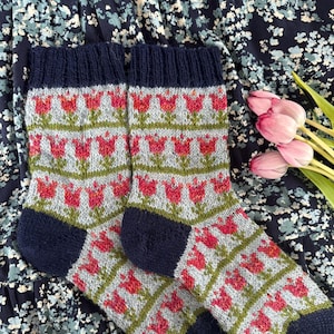 May include: A pair of knitted socks with a floral pattern. The socks feature a light gray base with red tulip designs, green stems, and navy blue cuffs and toes. The socks are displayed on a floral patterned background.