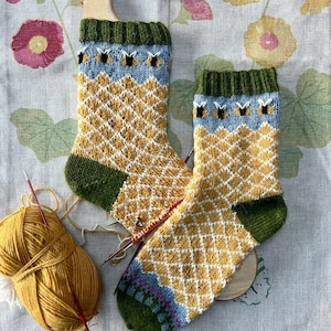May include: A pair of hand-knitted socks with a honeycomb pattern in yellow and white, green and blue accents, and a bee motif. The socks are displayed on a wooden sock blocker.