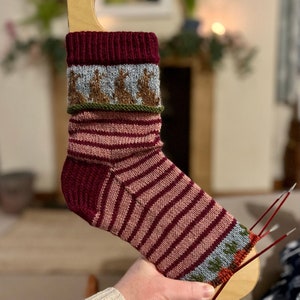 May include: A close-up of a knitted sock with a striped pattern in shades of pink and burgundy. The sock has a decorative band with a pattern of small brown figures on a light blue background. The sock is being held on a wooden sock blocker.