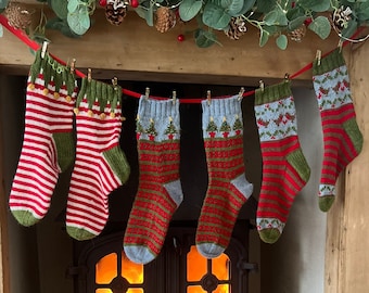 Peut inclure: Cinq chaussettes de Noël en tricot sont accrochées à une cheminée décorée d'une guirlande de verdure. Les chaussettes sont rouges, vertes et blanches avec différents motifs, notamment des arbres et des flocons de neige. Un poêle à bois est visible sous la cheminée, avec des flammes qui brûlent à l'intérieur. Un panier en osier rempli de laine se trouve sur le sol à droite du poêle.