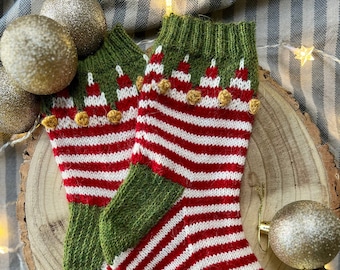 Peut inclure: Une paire de chaussettes de Noël tricotées à rayures rouges et blanches, avec des talons et des orteils verts, et des poignets verts avec des accents blancs et des pompons dorés. Les chaussettes sont sur une tranche de bois avec des boules de Noël dorées.
