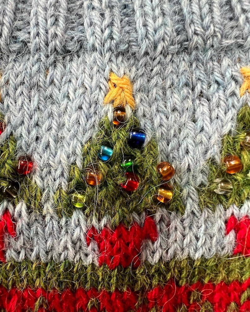 Peut inclure: Gros plan sur un motif de No&euml;l tricot&eacute;. Le tissu en tricot bleu clair pr&eacute;sente des sapins de No&euml;l verts brod&eacute;s orn&eacute;s de perles color&eacute;es. Un motif rouge et blanc se trouve en bas, et une &eacute;toile jaune surmonte chaque arbre.