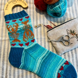 Pode incluir: Uma meia de tricô listrada azul-petróleo e azul-turquesa com um desenho de lagosta e corações vermelhos. A meia está pendurada em um gancho de madeira com o texto "From Devon with Love".