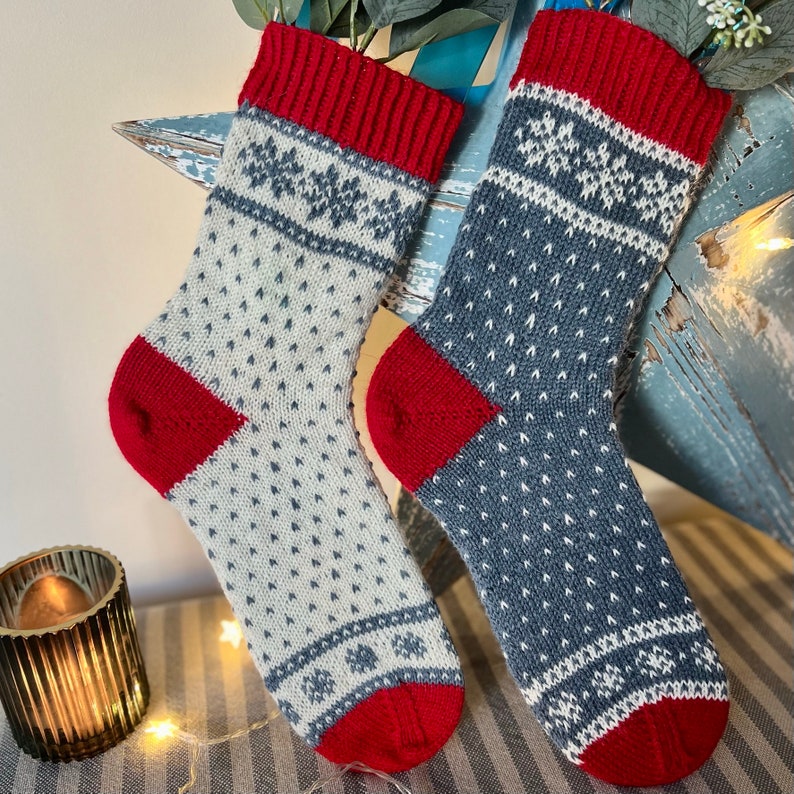 May include: Two pairs of knitted socks with a white and grey snowflake pattern and red cuffs. The socks are displayed on a wooden surface.