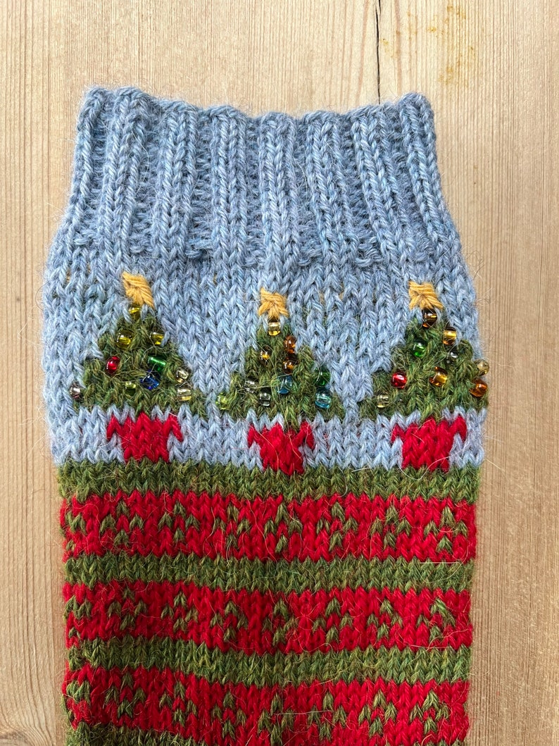 Peut inclure: Une chaussette de No&euml;l tricot&eacute;e avec un revers bleu clair et un corps &agrave; motifs rouges et verts. La chaussette pr&eacute;sente trois sapins de No&euml;l verts avec des &eacute;toiles dor&eacute;es et des ornements perl&eacute;s.