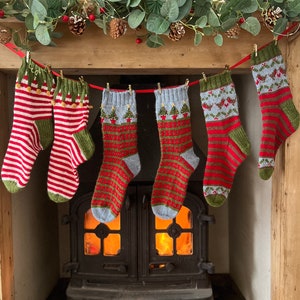 Peut inclure: Six chaussettes de No&euml;l tricot&eacute;es &agrave; la main, rouges, vertes et blanches, sont suspendues &agrave; un ruban rouge. Les chaussettes pr&eacute;sentent divers motifs, notamment des rayures, des arbres et des flocons de neige. Un foyer avec un feu allum&eacute; est visible en dessous.