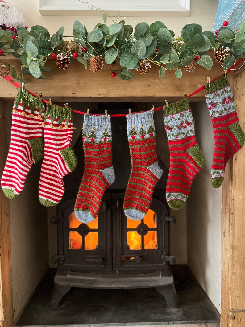Peut inclure: Six chaussettes de No&euml;l tricot&eacute;es &agrave; la main, rouges, vertes et blanches, sont suspendues &agrave; un ruban rouge. Les chaussettes pr&eacute;sentent divers motifs, notamment des rayures, des arbres et des flocons de neige. Un foyer avec un feu allum&eacute; est visible en dessous.