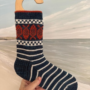 Pode incluir: Uma meia de lã tricotada à mão com um padrão de riscas azul-marinho e branco, uma faixa com detalhes axadrezados brancos e azul-marinho e uma fila de motivos de peixes cor de laranja ferrugem. A meia pende de um cabide de madeira gravado From Devon With Love.