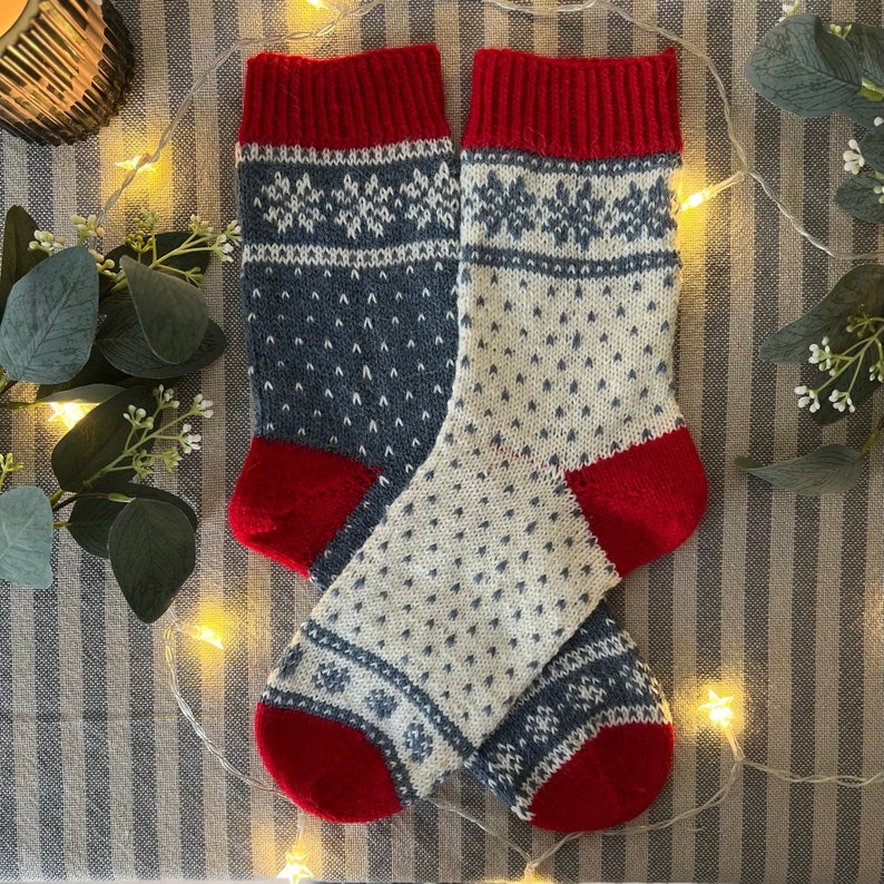 May include: A pair of knitted socks with a white and blue snowflake pattern and red cuffs. The socks are on a striped fabric background.