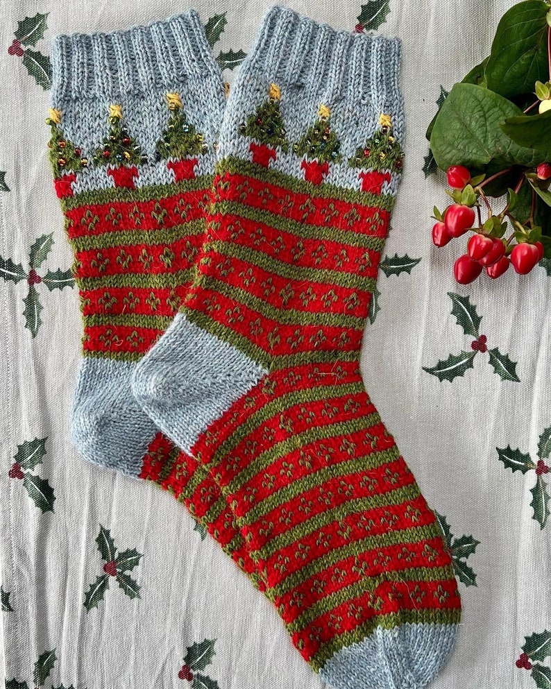 Peut inclure: Une paire de chaussettes de No&euml;l tricot&eacute;es. Les chaussettes pr&eacute;sentent un bord et un bout du pied bleu clair, avec un motif ray&eacute; rouge et vert sur le corps. Le bord pr&eacute;sente un motif de sapins de No&euml;l en pots.