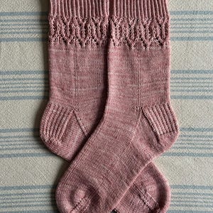 Könnte beinhalten: Ein Paar handgestrickte Socken in einem staubigen Rosaton. Die Socken haben ein dekoratives Lochmuster am Bündchen und Rippendetails an Ferse und Zehen. Die Socken liegen auf einem gestreiften Stoffhintergrund.